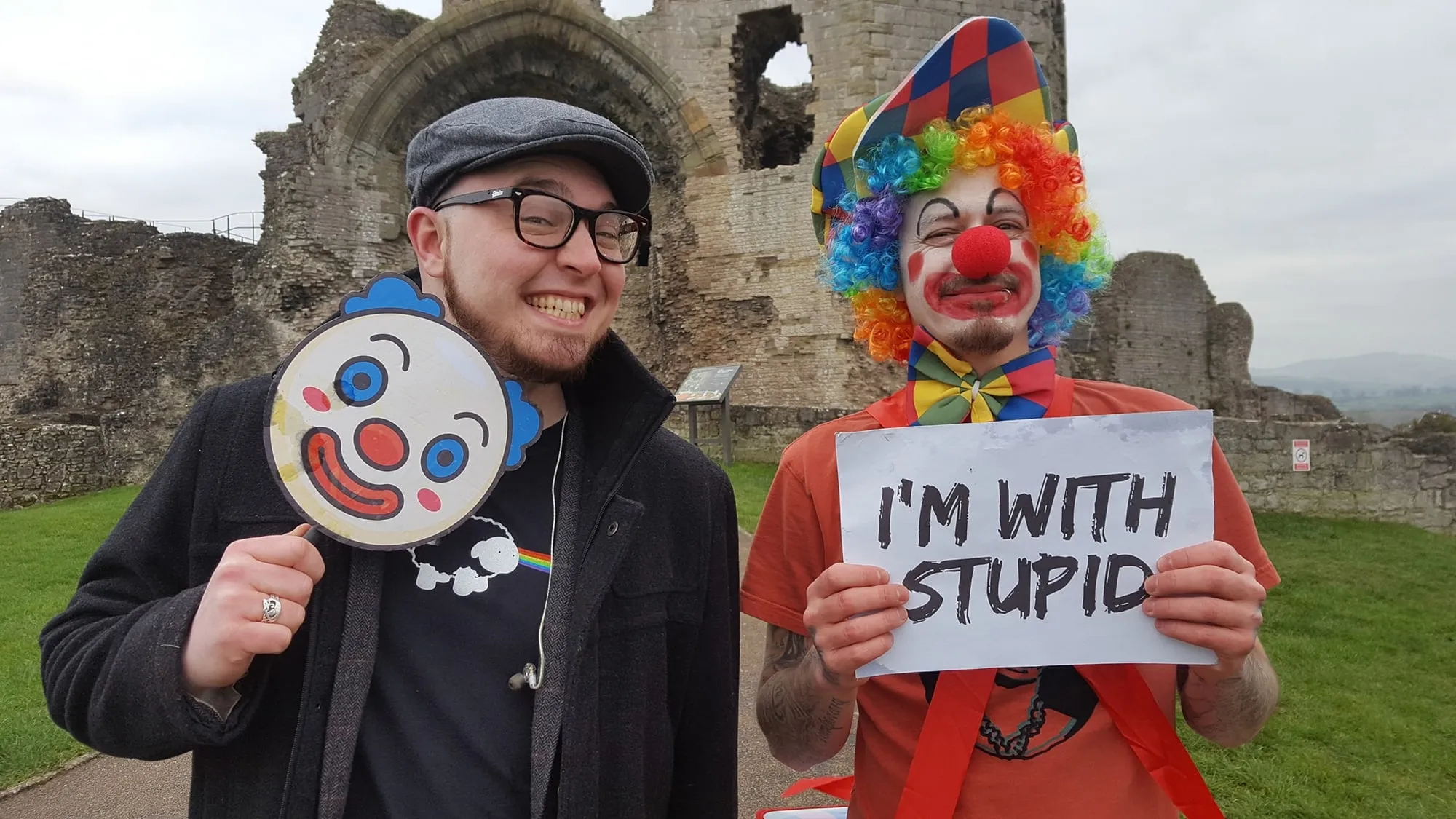 Isaac Birchall holding a clown mask and Andy Cooper dressed as a clown holding an I'm With Stupid sign, at Denbigh Castle during the A Certain Kind video shoot, 2019.